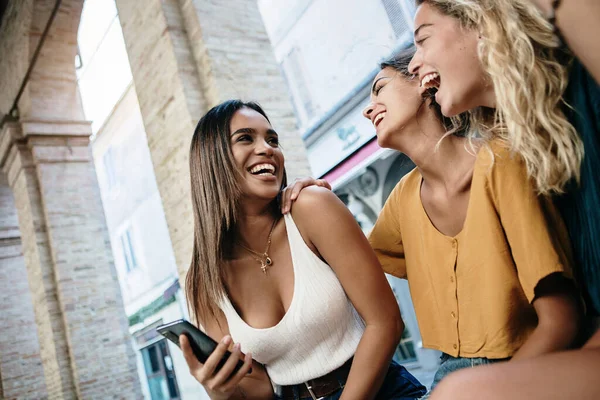 Three happy female friends laughing and having fun after watching social media content on mobile phone sitting outdoor - Multiracial young women enjoying free time together in city street