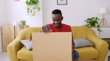Mutlu Afrikalı adam evdeki karton kutuyu açıyor genç milenyum erkeği kanepede oturuyor yeni kıyafetlerin paketini açıyor online alışveriş konsepti