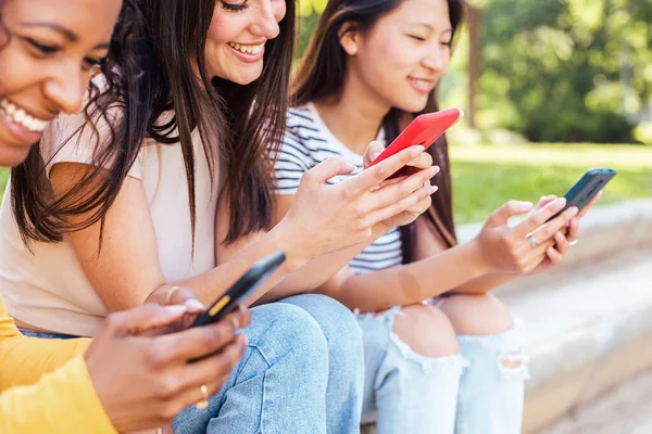 Dışarıda akıllı cep telefonu kullanan mutlu, genç, çok ırklı bayan arkadaşlar yazın dışarıda otururken elinde akıllı telefon tutan üç bin yıllık çeşitli kadın.