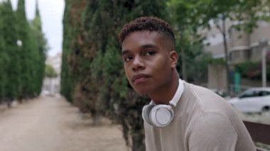 Dışarıda bankta oturan kulaklıklı Afro-Amerikalı genç bir adam. Milenyum öğrencisi çocuk parkta birini bekliyor. Yüksek kalite 4k görüntü