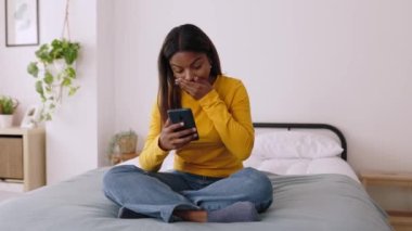 Excited african american young girl using mobile phone celebrating success at home. Cheerful millennial student woman reading good news on cellphone while sitting on bed. High quality 4k footage