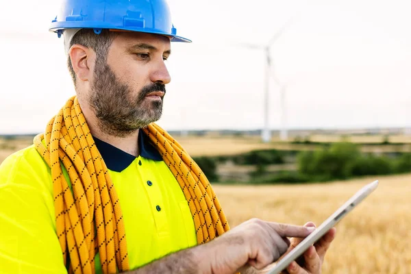Young technician worker man with blue safety helmet and climbing equipment working with digital tablet on wind turbine farm - Renewable energy concept