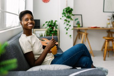 Genç Afrikalı bir kadın kanepede oturup portakal suyu içerken Millennial rüya gibi bir kadın evde dinlenirken modern dairede rahat bir koltukta oturuyor.