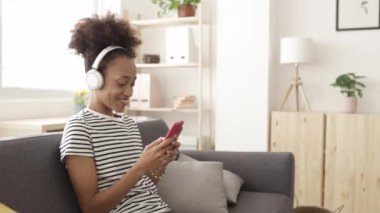Genç Afro-Amerikalı kız, evde koltukta otururken akıllı telefondan sohbet ediyor - Milenyum kadını cep telefonu uygulamasında sosyal medya içeriğini izlerken eğleniyor - teknoloji ve gençlik kavramı
