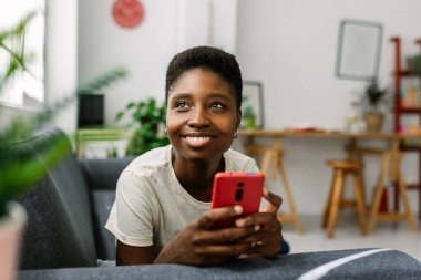 Güzel, genç Afrikalı bir kadın cep telefonuyla koltukta dinleniyor mutlu, rahat, akıllı bir kadın modern apartmandaki koltukta uzanmış akıllı bir telefon tutuyor.