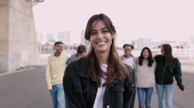 Şehirli arka planda bir grup çok ırklı arkadaşla dışarıda yürürken kameraya bakan mutlu, kendine güvenen genç bir kadının portresi. Takım çalışması, insan kaynakları ve topluluk üyeleri