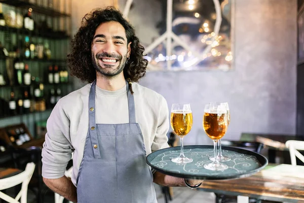 Friendly portrait of confident young adult waiter male holding serving tray with beers smiling at camera at brewery bar