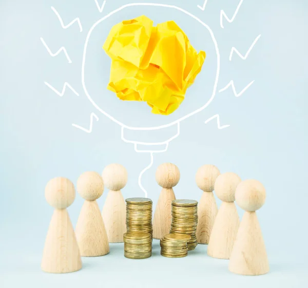 Business concept. Team concept and idea creation. Wooden figures of people stand around coins and a lamp on a blue background. Teamwork. Brainstorming Business Recovery and Growth. Copy space.