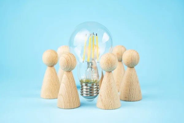 Business concept. Team concept and idea creation. Wooden figures of people stand around coins and a lamp on a blue background. Teamwork. Brainstorming Business Recovery and Growth. Copy space.