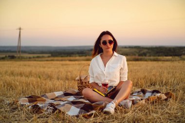 Mutlu esmer kız sonbahar günbatımında buğday tarlasında piknik yaparken şarap içiyor.