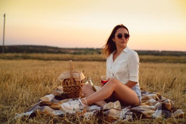 Mutlu esmer kız sonbahar günbatımında buğday tarlasında piknik yaparken şarap içiyor.