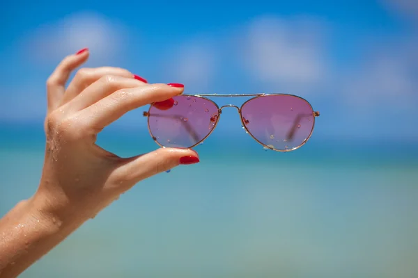 Womans hand holding sunglasses on tropical beach — Stock Photo, Image