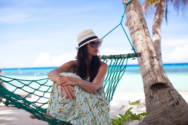 Kvinna i hatt sitter i hängmatta i trädets skugga på en strand — Stockfoto