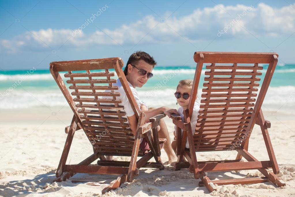 Young father and his wonderful daughter sitting on beach wooden chairs looking at camera