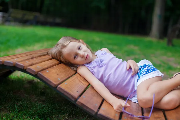 Roztomilá holčička v purpurové narozeninám brýle ležící na venkovní dřevěné židle — Stock fotografie
