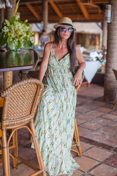 Beautiful brunette woman in long dress and hat relaxing near bar in exotic resort — Stock Photo, Image