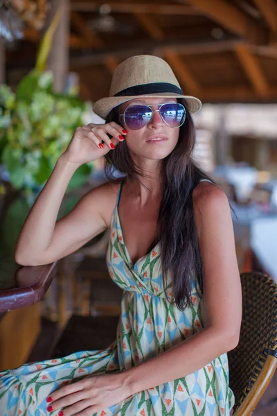 Beautiful brunette woman in long dress and hat relaxing near bar in exotic resort — Stock Photo, Image