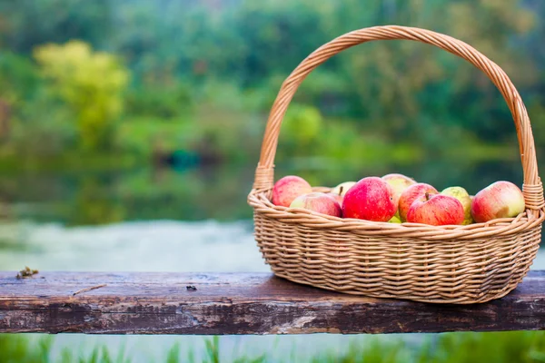 Big straw basket with red and yellow apples on a bench by the lake — Stock Photo, Image