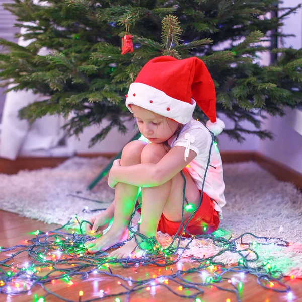 Bambina bellissima in cappello Babbo Natale seduta sotto l'albero di Natale tra ghirlande — Foto Stock