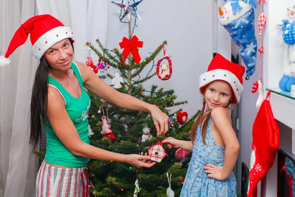 Junge Mutter und ihr entzückendes kleines Mädchen in Weihnachtsmannhüten schmücken ihren Weihnachtsbaum — Stockfoto