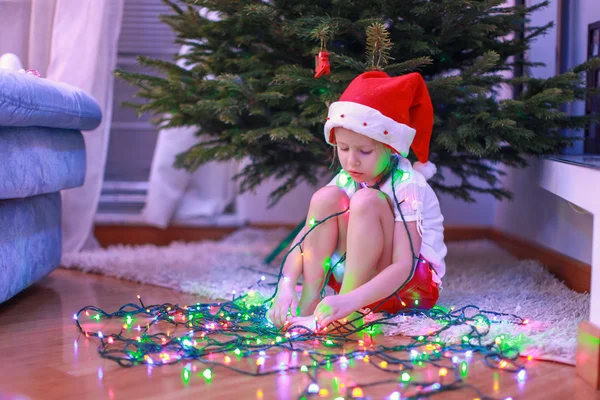 Little beautiful girl in Santa Claus hat sitting under the Christmas tree among garlands — Stock Photo, Image