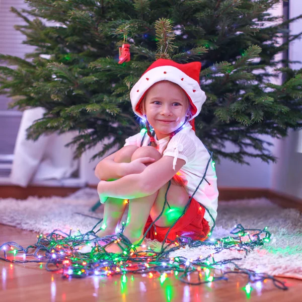 Pequena menina bonita em chapéu de Papai Noel sentado sob a árvore de Natal entre grinaldas — Fotografia de Stock