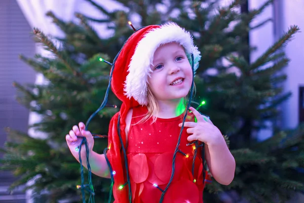 Pequena menina bonita em um vestido vermelho e chapéu com guirlandas de Natal em torno de seu pescoço — Fotografia de Stock