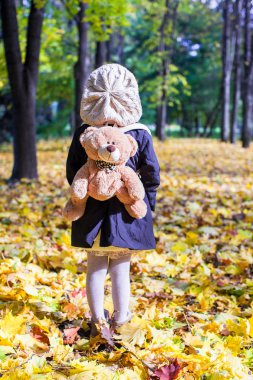 Rear view of charming little girl with a backpack-bear behind in the autumn sunny forest