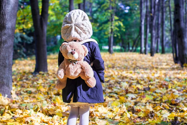 Rear view of charming little girl with a backpack-bear behind in the autumn sunny forest — Stock Photo, Image