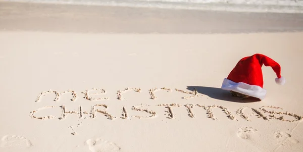 Kerstman hoed op wit zandstrand en merry christmas geschreven in het zand — Stockfoto