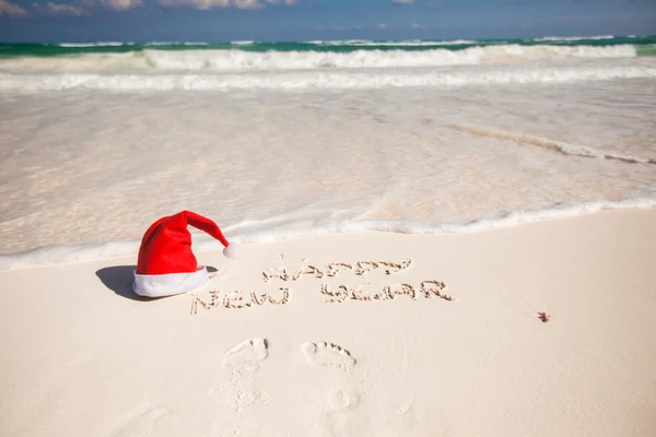 Santa kapelusz na białej, piaszczystej plaży i szczęśliwego nowego roku, napisany w piasku — Zdjęcie stockowe