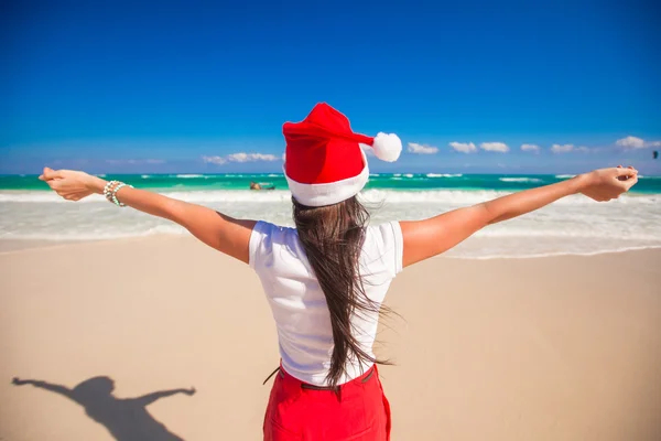 Achteraanzicht van mooi meisje in santa hat wandelen verspreiden haar handen op witte strand — Stockfoto