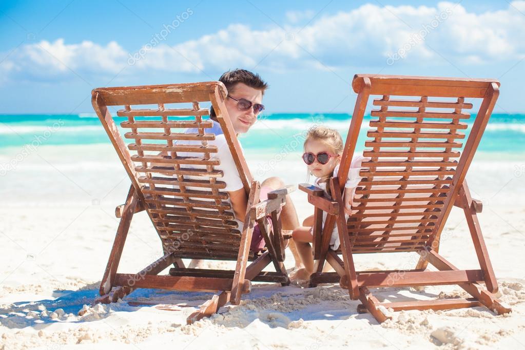 Little cute girl with her young dad sitting on beach wooden chairs looking at camera