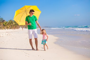 genç Baba ve küçük kızı beyaz kum plajı üzerinde sarı bir şemsiye altında yürüyüş