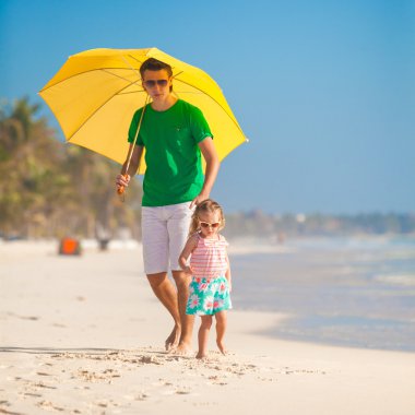 genç Baba ve küçük kızı beyaz kum plajı üzerinde sarı bir şemsiye altında yürüyüş