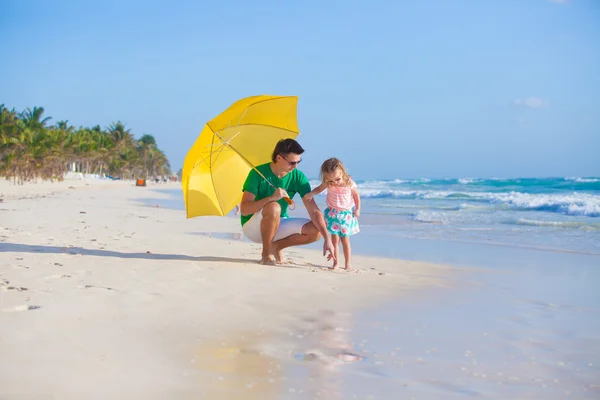 Young father and his adorable little daughter hiding from the sun under a yellow umbrella on white sunny day — Stock Photo, Image