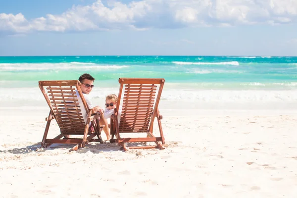 她年轻的父亲坐在沙滩木椅看着摄像机上的小女儿 — 图库照片