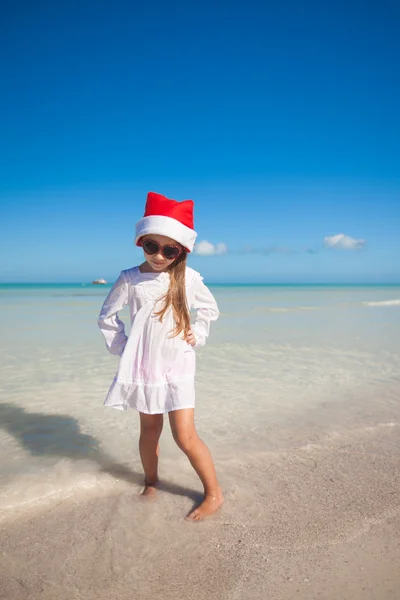 Pequena menina bonito em chapéu vermelho Papai Noel e óculos de sol na praia — Fotografia de Stock