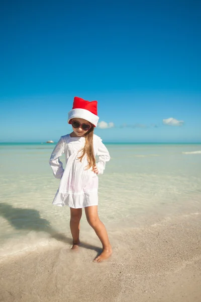 Beetje schattig meisje in de rode hoed santa claus en zonnebril op het strand — Stockfoto