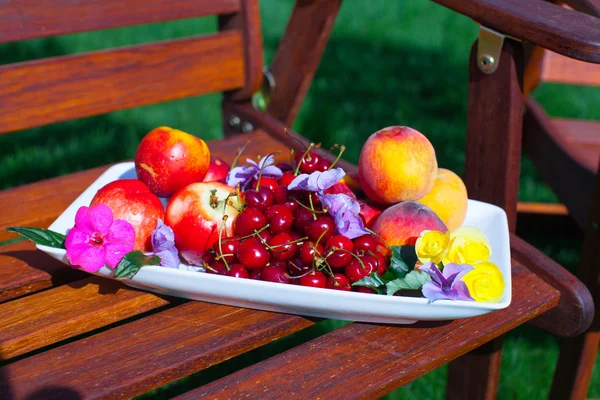 Тарелка со свежими фруктами и цветами на деревянных стульях в саду — стоковое фото