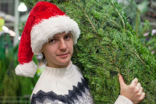 Portrét mladého muže v santa hat koupit vánoční stromek a a ukazuje palec — Stock fotografie