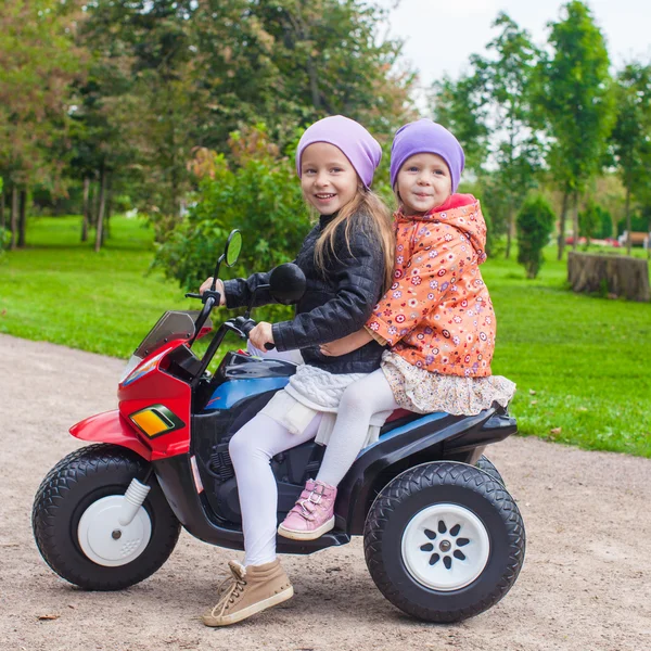 Dos pequeñas y hermosas hermanas sentadas en una motocicleta de juguete en un parque verde —  Fotos de Stock