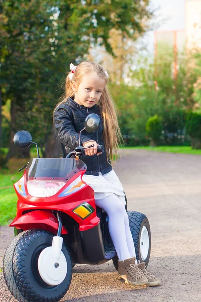Adorable happy little girl in leather jacket sitting on her toy motorcycle outdoors — Stock Photo, Image