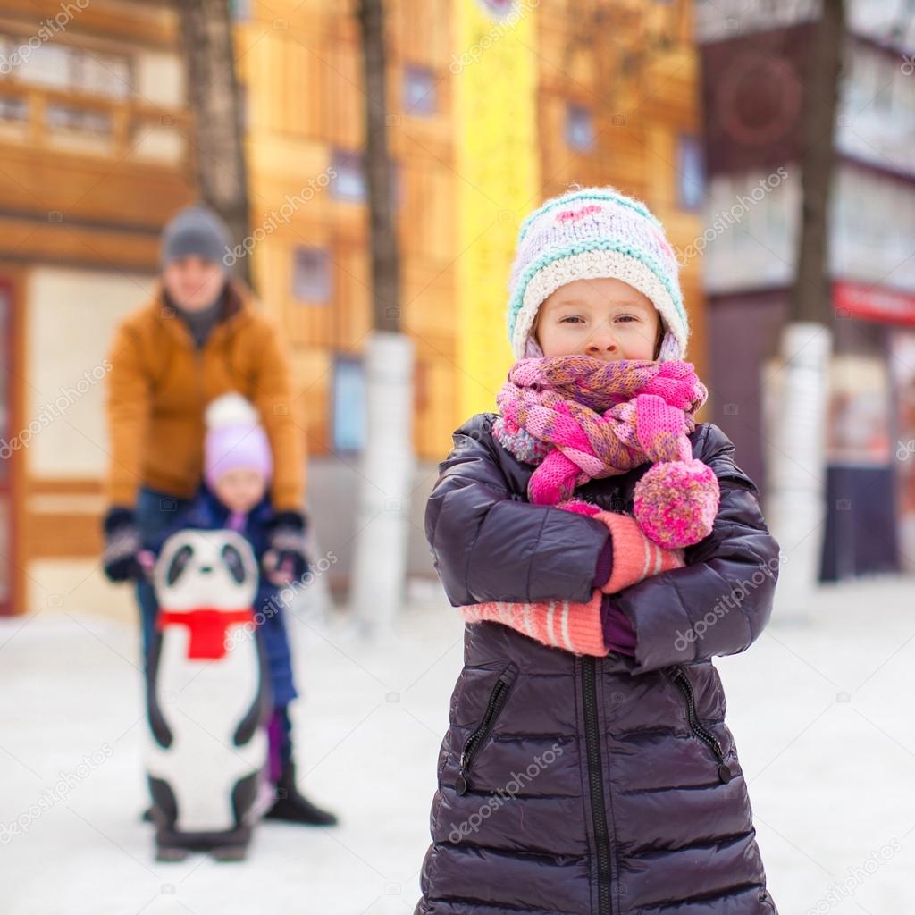 Adorable girl on skating rink, dad with little sister in the background