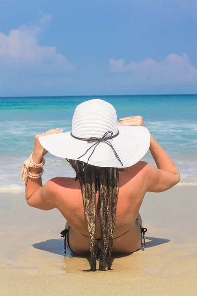 Vue arrière de la jeune femme assise dans un chapeau sur une plage de sable blanc profitant de vacances d'été regardant l'océan . — Photo