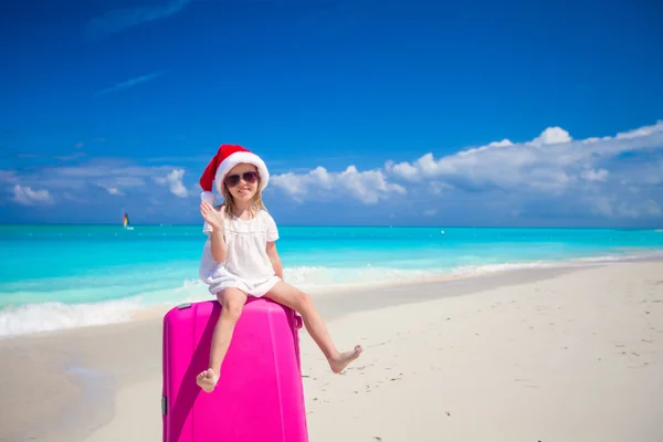 Bambina in cappello Babbo Natale seduta su una grande valigia sulla spiaggia tropicale — Foto Stock