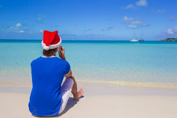 Vista posteriore del giovane in cappello di Babbo Natale con telefono sulla spiaggia di caraibi tropicali — Foto Stock