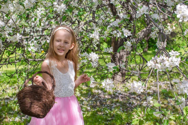 Bedårande liten flicka med halm korg i blommande äppelträdgård — Stockfoto