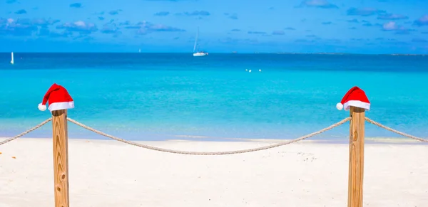 Primo piano due cappelli di Babbo Natale sulla recinzione alla spiaggia bianca tropicale — Foto Stock