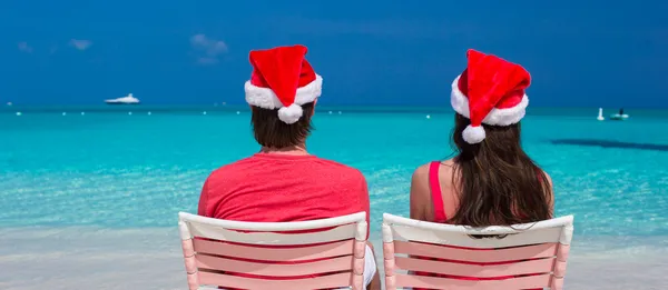 Rückansicht eines jungen Paares in roten Weihnachtsmützen, das auf Strandstühlen sitzt — Stockfoto